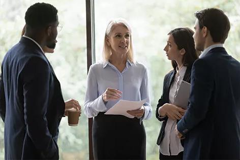 A group of people in business attire gather near a bright window to chat.